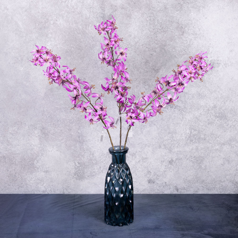 Three faux lily stems in a deep pinkish purple colour, shown in a blue glass vase