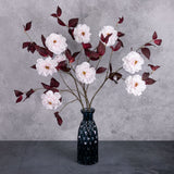 A group of three faux camelia sprays, in white with red leaves, displayed in a blue glass vase