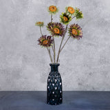 A group of three faux sunflower sprays in a muted purple colour, with tinges of green, showing three flowers, displayed in a blue glass vase