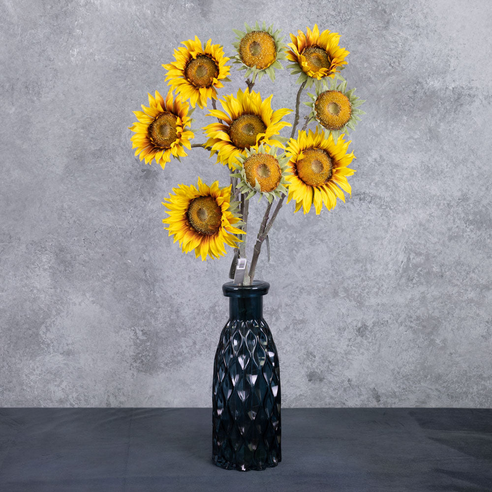 A group of three faux sunflower sprays in a rich yellow colour, displayed in a blue glass vase