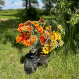 A group of different coloured, sized, and shaped faux sunflowers, displayed in a meadow against a bright, sunny sky