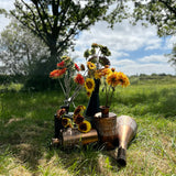 A group of different coloured, sized, and shaped faux sunflowers, displayed in a meadow against a bright, sunny sky